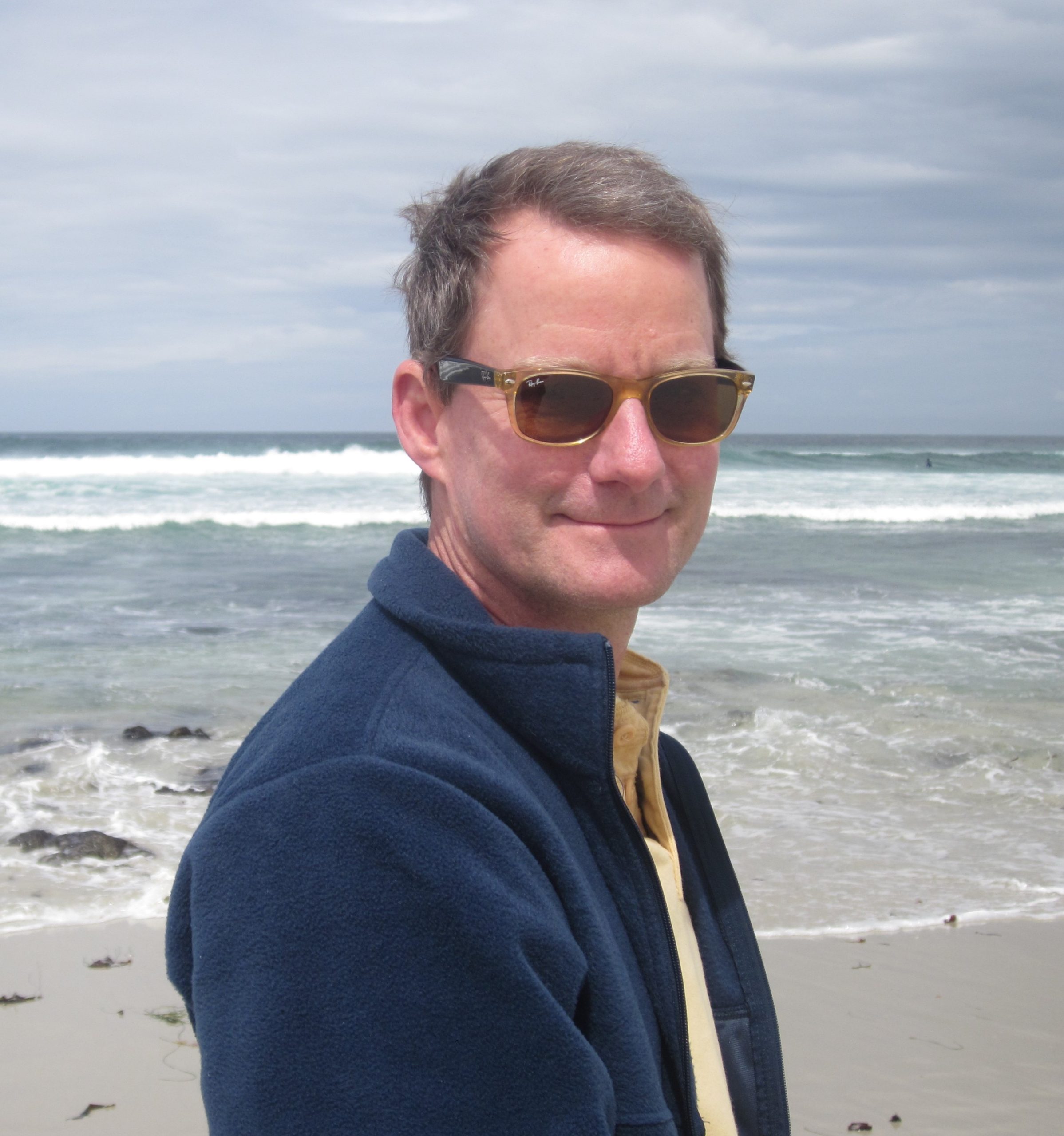 Guy Peters in sunglasses stands confidently on a sunny beach, savoring the coastal view.